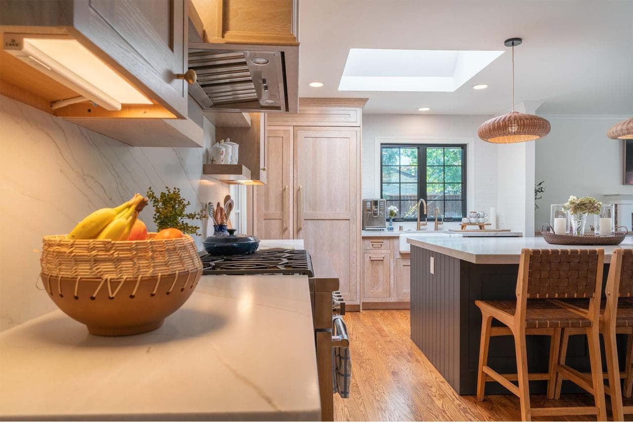 After: Open-concept kitchen remodel by ASL Remodeling featuring white oak cabinets, quartz waterfall island with navy base, skylight, rattan pendants, and integrated paneled refrigerator
