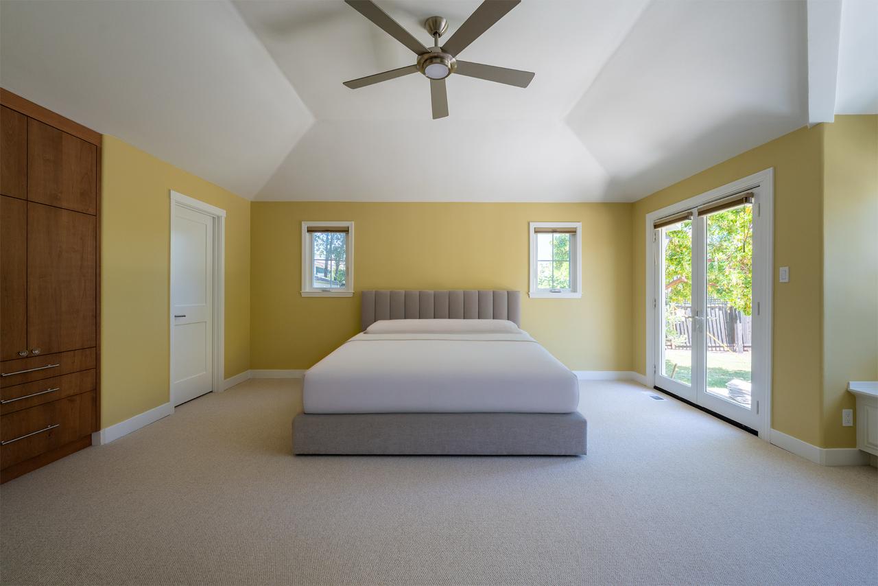 Bright second story addition bedroom in Los Gatos featuring bold yellow accent wall, modern ceiling fan, and large windows providing abundant natural light.